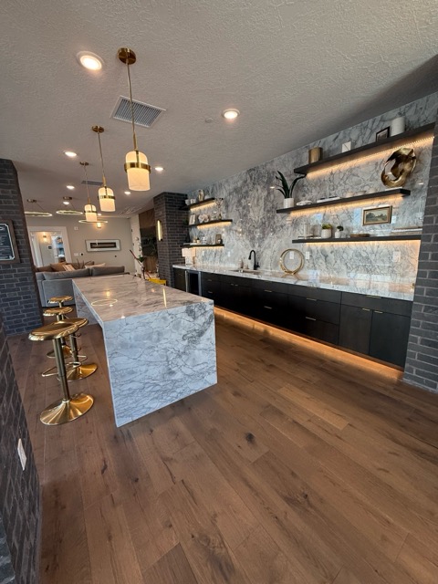 Clubhouse bar with a marble waterfall island, matte black slab cabinetry and LED-lit floating shelves against charcoal brick
