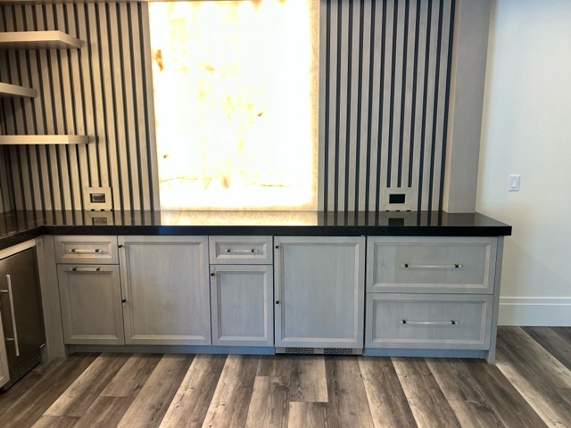 Driftwood-stained shaker bar cabinets with acrylic bar pulls, black quartz countertop and a full-height backlit onyx panel