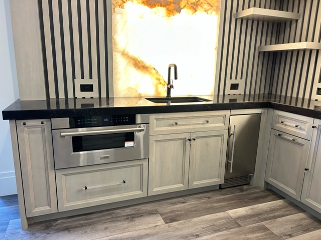 Wet bar sink station with a matte black pull-down faucet, stainless microwave drawer and gold-veined backlit onyx backsplash