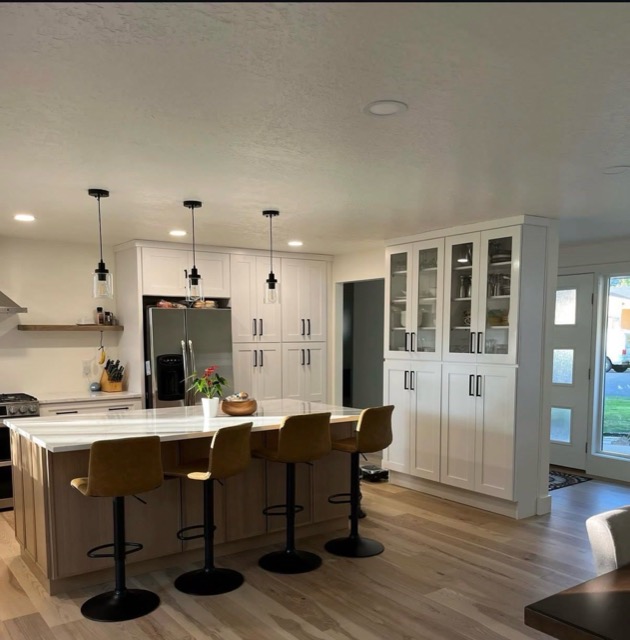 View across a white-oak kitchen island with black pendant lights toward a glass-front china hutch