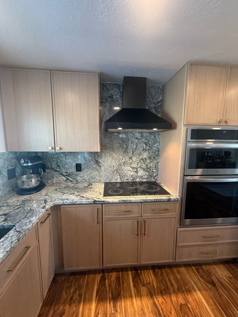 Pale rift-oak cabinets with brass hardware, a black wall-mount chimney hood and full-height grey-green granite backsplash